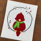 An applique of a cute plump cardinal wearing a scarf and sitting on a twig wreath with berries by snugglepuppyapplique.com