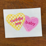 An applique of candy hearts with the words "cuddle me" and "baby" embroidered on them by snugglepuppyapplique.com
