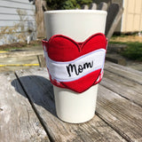 A cup cozy with a heart and scroll with the word "Mom" embroidered on it by snugglepuppyapplique.com