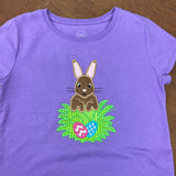 An applique on a purple shirt of a bunny rabbit discovering two eggs in the grass by snugglepuppyapplique.com