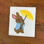 An applique embroidery design of a rabbit in a coat holding an open umbrella by snugglepuppyapplique.com