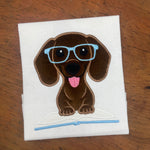 An applique of a dachshund wearing glasses and his paws on an open book by snugglepuppyapplique.com