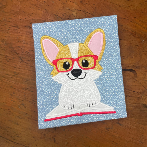 An appliqué of a corgi wearing glasses with his paws on an open book by snugglepuppyapplique.com
