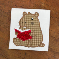 An applique of a capybara reading a book by snugglepuppyapplique.com