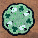 A felt candle mat with sheep holding clover in their mouths and large and small clover in a scalloped circle. by snugglepuppyapplique.coom