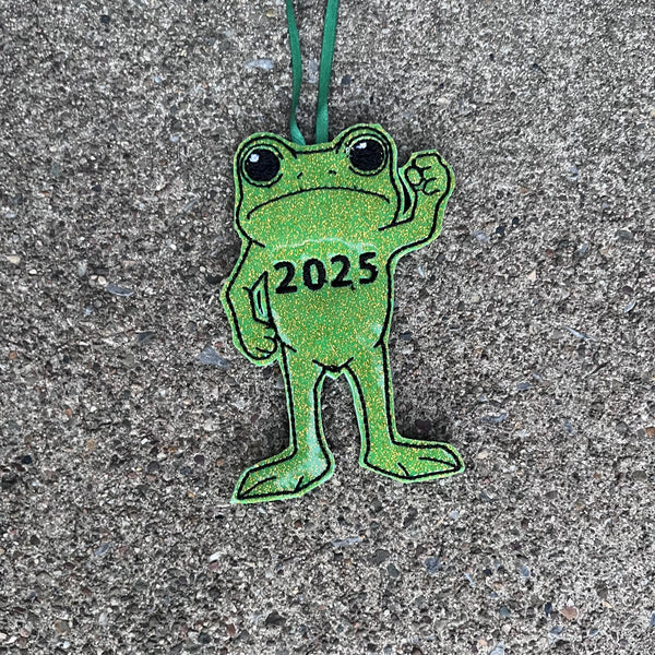 An ITH Christmas ornament of a Frog with his fist in the air in protest and "2025" embroidered on his chest.  by Snugglepuppyapplique.com