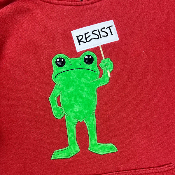 Red sweatshirt with an applique of a green frog holding a 'RESIST' sign on a red background