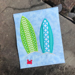An applique of two surfboards stuck in the sand and an embroidered crab, by snugglepuppyapplique.com