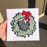An applique of a cat with its head through a christmas wreath batting at the bow by snugglepuppyapplique.com