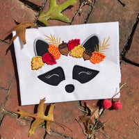 An applique of a raccoon's face wearing a crown of fall leaves and acorns by snugglpuppyapplique.com