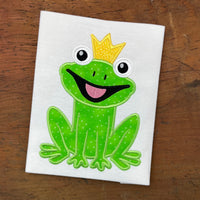 An applique of a smiling frog wearing a crown by snugglepuppyapplique.com