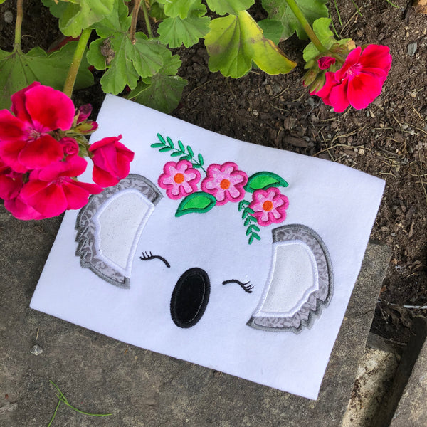 an applique design of a koala's ears, nose and closed eyes with lashes wearing a crown of flowers and leaves by snugglepuppyapplique.com