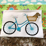 An applique of a bicycle with a basket and a dachshund inside the basket. snuggleppyapplique.com